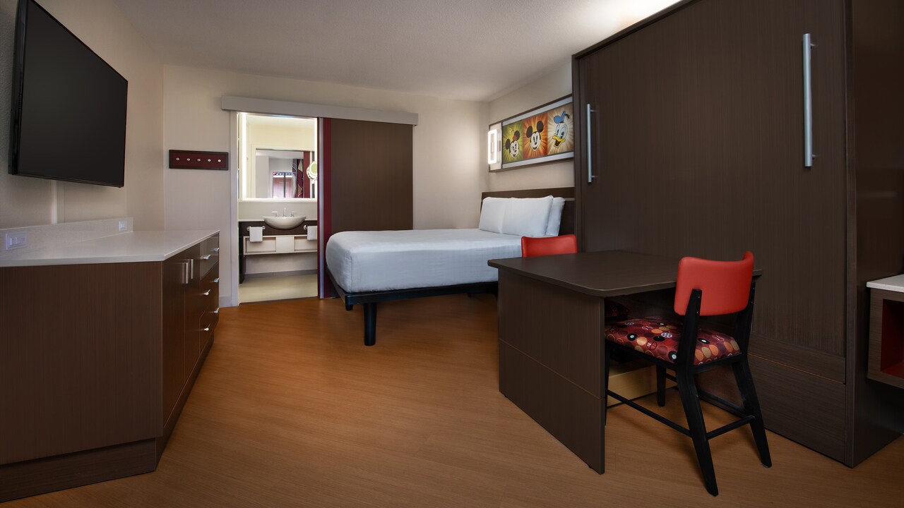 A table with a chair and a dresser across from a bed with a night stand
