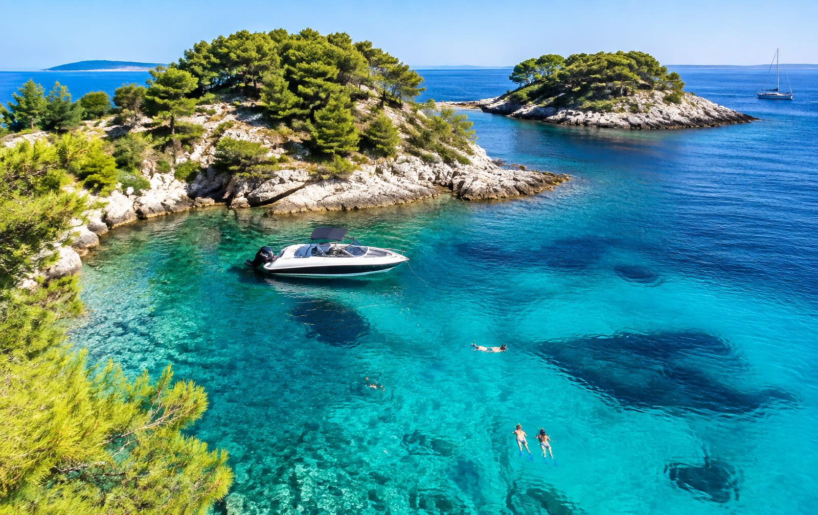 A speedboat anchored in a clear turquoise cove among the pine-covered Pakleni Islands, with swimmers snorkeling