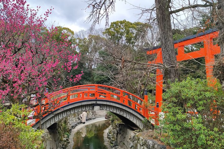 Vermilion arched bridge over a stream, with pink blossoms on the left and a torii on the right