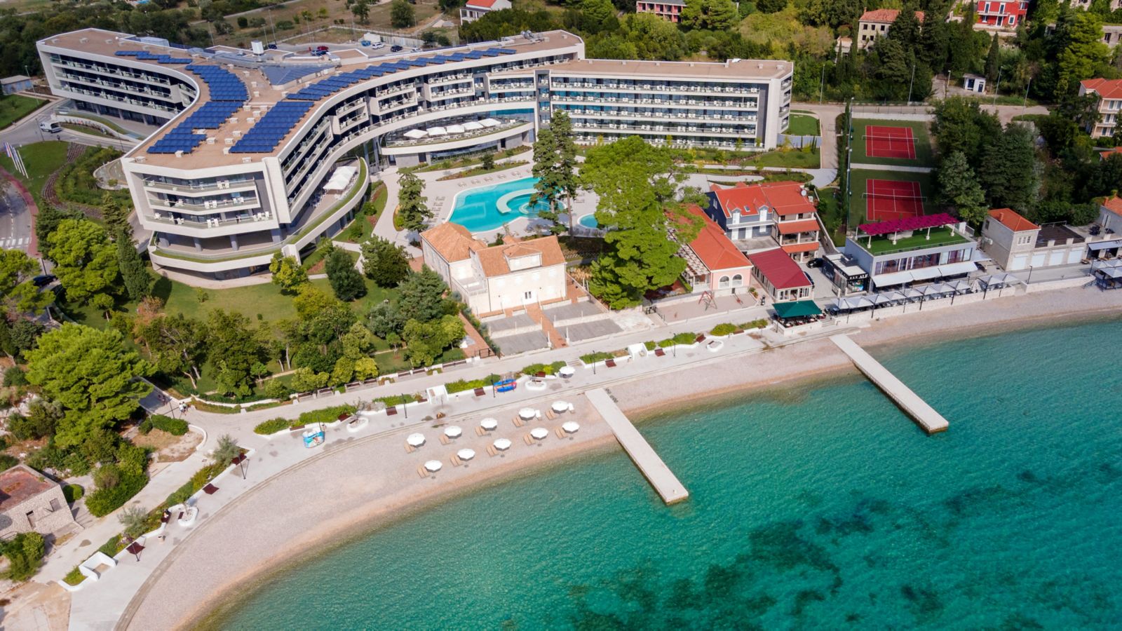 Aerial of Sheraton Dubrovnik Riviera, with the curved hotel, outdoor pool, two tennis courts, and the pebble beach