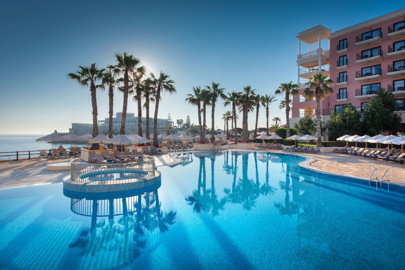 Bay pool with palm trees, white umbrellas, and the sea beside the hotel